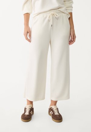 Persoon die witte wijde cropped broek met trekkoord taille en bruine sneakers met witte veters en strepen draagt, staande tegen een effen achtergrond.
