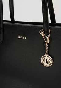 Czarna torebka z ekoskóry z złotym logo DKNY, podwójnymi uchwytami i złotym zawieszką przymocowaną łańcuszkiem. Gładka faktura o minimalistycznym designie.