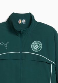 Dunkelgrüne Zip-Jacke mit hohem Kragen, ausgestattet mit einem weißen Streifen, Puma-Logo und dem Vereinswappen von Manchester City auf der linken Seite.