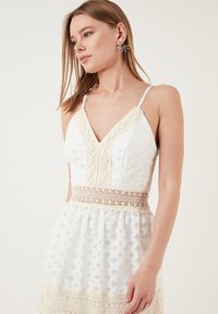 Weißes Kleid mit verstellbaren Spaghetti-Trägern, V-Ausschnitt, Spitzenbesatz, strukturiertem Stoff und Häkeldetails an der Taille und dem Saum.