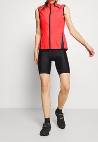 Rød ærmeløs cykelvest med høj krave, med en frontlynlås, sorte sideakcenter og en strømlinet pasform. Båret sammen med sorte shorts.