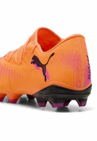 Oranžs futbola apavu modelis ar teksturētu sintētisko materiālu, rozā akcentiem, melnu zoli un izteiksmīgu melnu kaķa logotipu sānos.