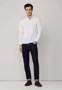 Bijela polo majica s dugim rukavima i okovratnikom na kopčanje, u kombinaciji s tamnoplavim trapericama uskog kroja i smeđim cipelama, s podvrnutim rukavima.