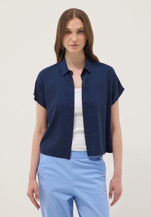 Jeune femme aux cheveux bruns lisses porte une chemise bleu marine à manches courtes boutonnée sur un débardeur blanc et un pantalon bleu clair, debout devant un fond blanc.