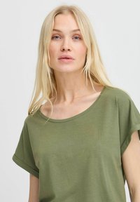Camiseta verde oliva con un escote amplio y mangas cortas enrolladas, hecha de una tela suave y ligera. Textura lisa, ajuste casual.