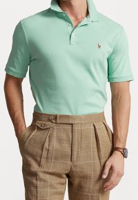 Light mint green polo shirt with a two-button placket and small embroidered logo, paired with brown plaid high-waisted trousers.
