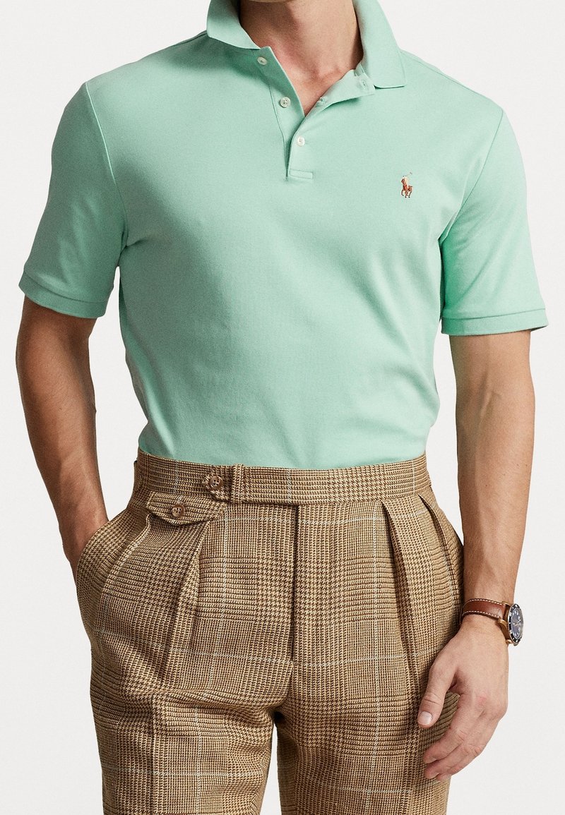 Light mint green polo shirt with a two-button placket and small embroidered logo, paired with brown plaid high-waisted trousers.