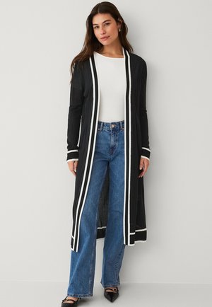 Cardigan long noir à rayures blanches, en matériau léger, avec un design ouvert à l'avant, porté par-dessus un haut blanc et associé à des jeans à jambe large bleus.