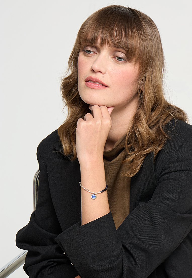 Silberarmband mit einem blauen Edelstein und einer Mischung aus dunklen Perlen. Das Model trägt ein schwarzes Blazer und ein braunes Oberteil, sitzt in einer nachdenklichen Pose.