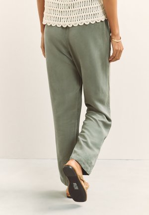 Personne portant un pantalon vert ample et un haut blanc en crochet, marchant loin avec des sandales marron sur un sol clair.