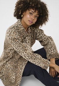 Srajca-jakna v leopardjem potisku v bež in rjavih odtenkih, ki se ponaša s sproščenim krojem, dolgimi rokavi in sprednjimi žepe. Temno jeans hlače so vidne spodaj.