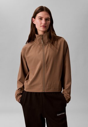 Femme portant une veste marron zippée à col haut et un pantalon Calvin Klein noir, debout les mains dans les poches devant un fond gris.