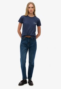 Marineblauwe katoenen t-shirt met een klein wit logo, gecombineerd met hoge, donkere spijkerbroek en zwarte nette schoenen.