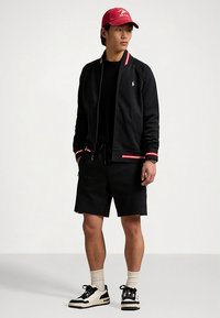 Juoda striukė su užtrauktuku, puošta raudonomis ir baltomis juostelėmis, derinama su juodais šortais ir juodai baltais sportbačiais. Raudona kepurė užbaigia aprangą.