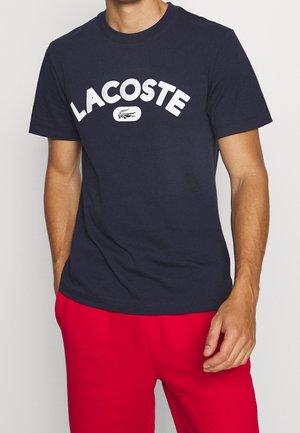 Granatowa bawełniana koszulka z odważnym białym logo "LACOSTE" i grafiką krokodyla, okrągłym dekoltem i krótkimi rękawami.