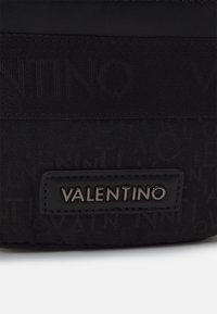 Svart Valentino-väska i texturerad tyg med upphöjt varumärkeslogo, stilren design och en slät läderdetalj. Metallisk hårdvarudetalj synlig.