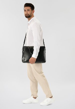 Borsa a tracolla in pelle nera con cerniere e manici, forma rettangolare, indossata da una persona con camicia bianca e pantaloni beige. Design semplice.