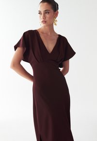 Burgundská šaty s hlbokým výstrihom do V, splývavými krátkymi rukávmi a štíhlym strihom. Disponujú bočným rozparkom a hladkou textúrou látky. Zlaté obľúbené náušnice.