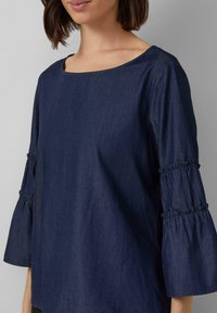 Dunkelblaues Denim-Oberteil mit weitem Ausschnitt, drei Viertel langen, ausgestellten Ärmeln mit gerafften Akzenten und einer glatten, strukturierten Oberfläche.