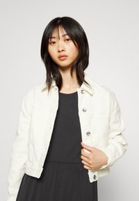 Jaqueta de ganga branca cropped com botões prateados, dois bolsos frontais e um tecido ligeiramente texturizado, usada sobre um vestido escuro.