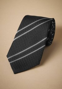 Cravatta nera in tessuto testurizzato con strisce bianche diagonali, caratterizzata da un motivo intrecciato. Arrotolata, mostrando un'estremità affusolata.