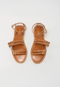 Sandales en cuir brun avec des sangles au niveau de la cheville, présentant deux sangles horizontales, des accents en métal et des coutures contrastées le long de la semelle.