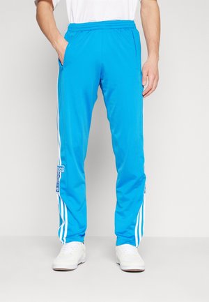 Man in heldere blauwe Adidas sportbroek met witte strepen aan de zijkanten en witte sneakers, staand tegen een effen muur.