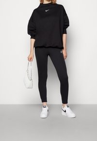 Svart sweatshirt med rund halsringning och logotyp, tillsammans med svarta figurnära leggings och vita sneakers. Håller en liten vit quiltad väska.