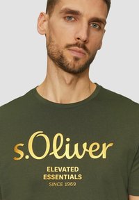 Mann mit kurzem braunem Haar und Bart, der ein dunkelgrünes T-Shirt mit goldenen Aufdruck "s.Oliver Elevated Essentials Since 1969" trägt.