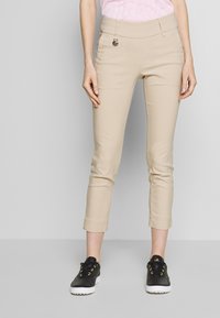 Beżowe spodnie 7/8 o gładkiej fakturze, z przednimi kieszeniami i srebrnym akcentem na pasku w talii, zestawione z czarnymi tenisówkami.