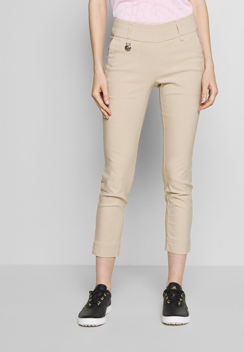 Beżowe spodnie 7/8 o gładkiej fakturze, z przednimi kieszeniami i srebrnym akcentem na pasku w talii, zestawione z czarnymi tenisówkami.