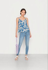 Top de alças azul floral com flores brancas, combinado com jeans skinny azul claro. A modelo segura uma bolsa de malha lavanda e usa sandálias brancas.
