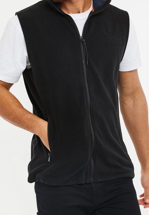 Homme portant une veste polaire noire zippée sur un t-shirt blanc à manches courtes, une main dans la poche de la veste, visible des épaules jusqu'aux hanches.