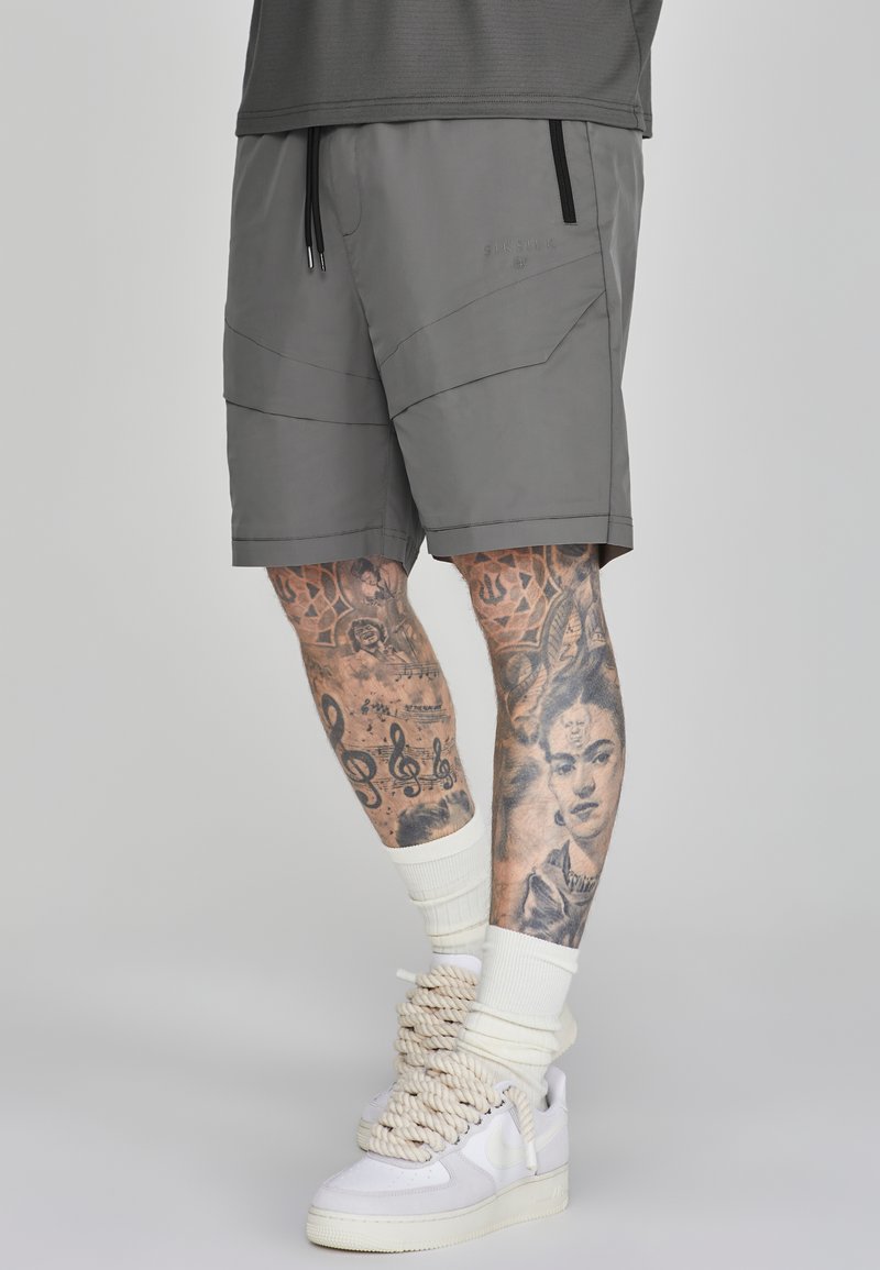 Grijze sportshorts met een trekkoord, voorzien van geometrische panelen en zijzakken. Draagt met witte hoge sneakers en gestippelde sokken.