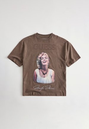 T-shirt marron à manches courtes avec le texte « GUESS », une femme souriante portant une robe blanche et un collier, et une signature sous l'image.