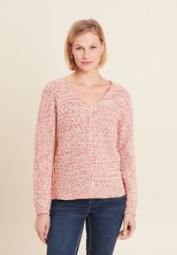 Rosa strukturierte Strickjacke mit V-Ausschnitt, die ein geometrisches Muster und lange Ärmel hat, kombiniert mit blauen Jeans.