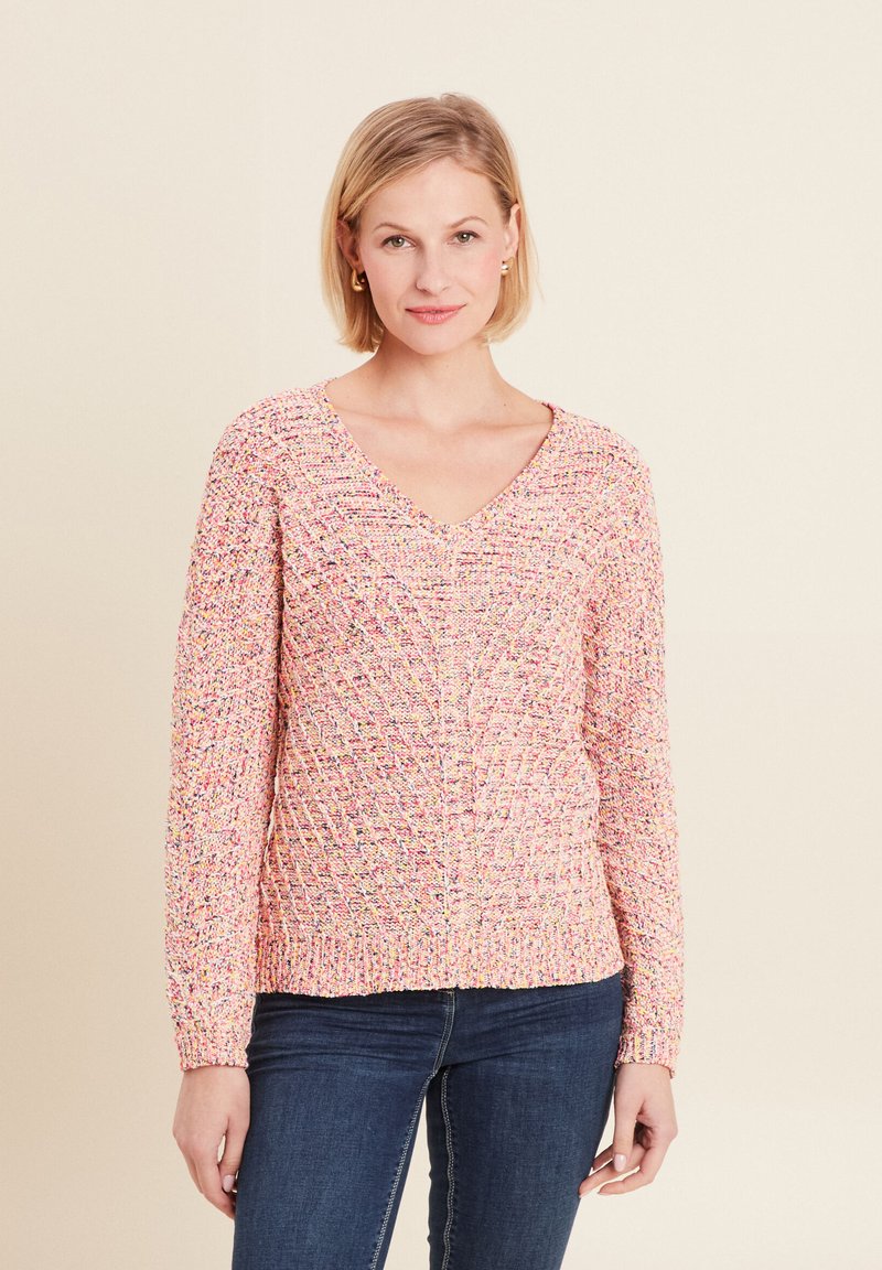 Rosa strukturierte Strickjacke mit V-Ausschnitt, die ein geometrisches Muster und lange Ärmel hat, kombiniert mit blauen Jeans.