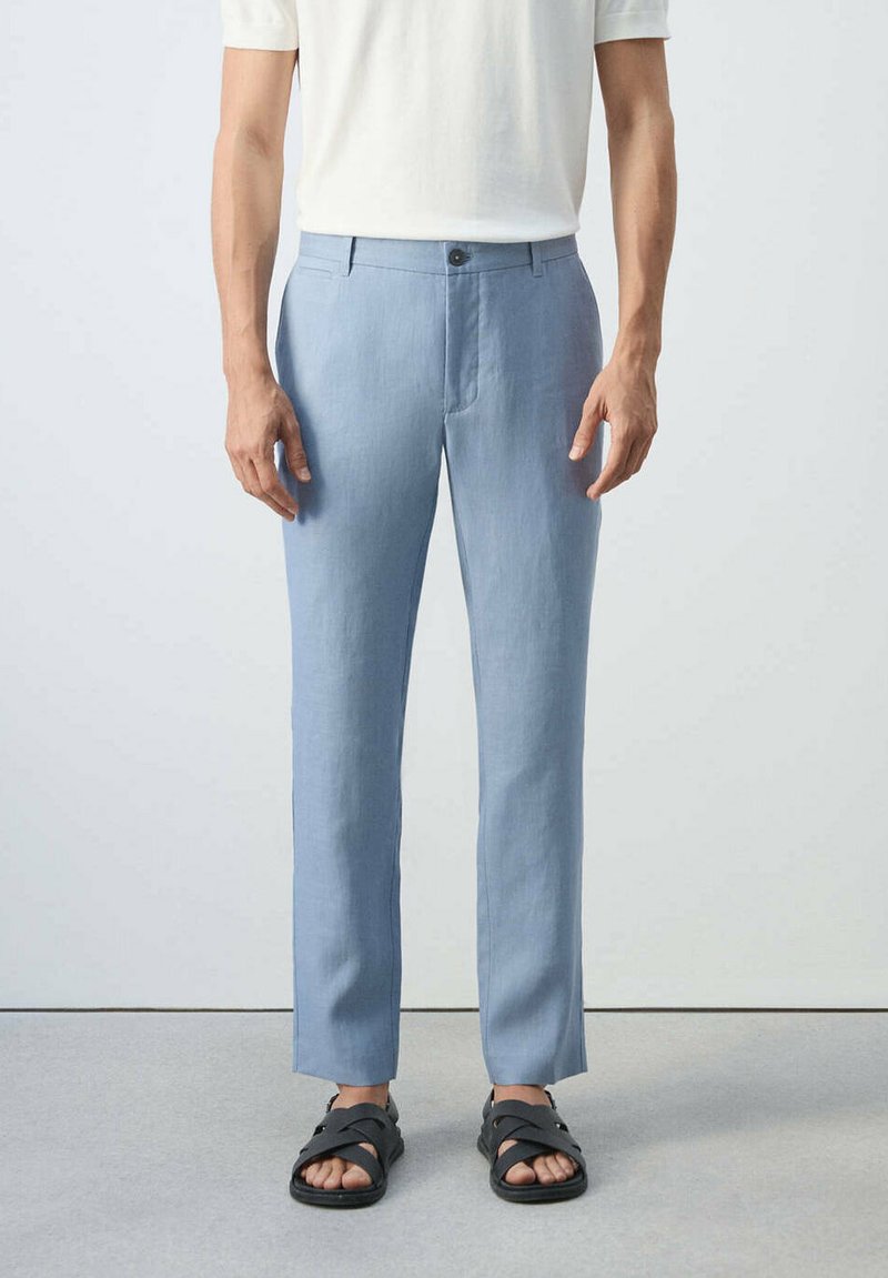 Un homme porte un pantalon bleu clair ajusté et des sandales noires à brides, assortis d'une chemise blanche à manches courtes, debout sur un sol gris.
