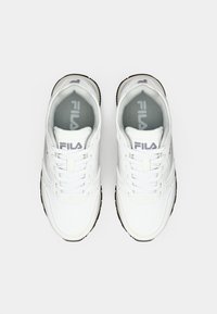 Bijele tenisice na vezivanje od kože s glatkom teksturom, sivi Fila logo i crna gumena potplat. Klasični sportski dizajn.