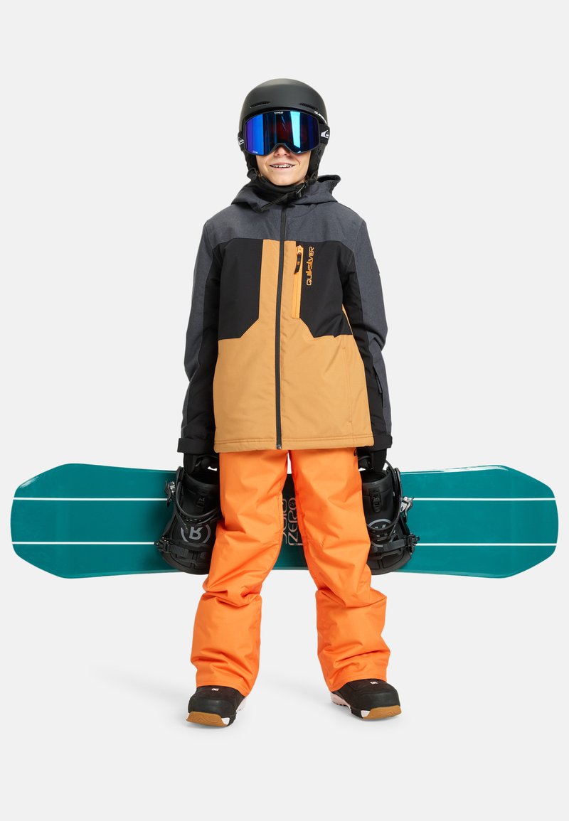 Ein Snowboarder trägt eine schwarze und orange Jacke, leuchtend orange Hosen und einen schwarzen Helm. Er hält ein türkisfarbenes Snowboard mit schwarzen Bindungen.