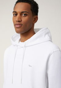 Hoodie blanc avec cordon de serrage, arborant un petit logo brodé. Le tissu semble lisse, avec une coupe décontractée et une texture douce.