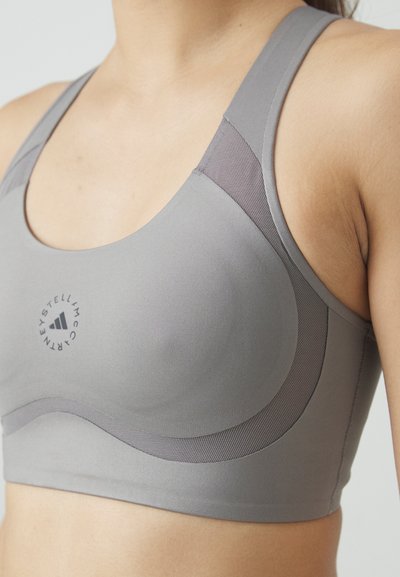 Sujetador deportivo gris con tela suave, diseño curvado, detalles de malla y logo. Cuenta con tirantes anchos y un acabado sin costuras para mayor comodidad.