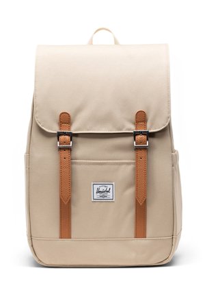 Beżowy plecak z tkaniny z dwoma jasnobrązowymi skórzanymi paskami na klapce przedniej oraz małą białą prostokątną etykietą marki poniżej.