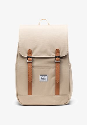 Bež platneni ruksak s dva kožna remena svijetlosmeđe boje s kopčama na prednjoj preklopnici i malom bijelom pravokutnom oznakom marke ispod.