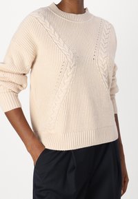 Pull en tricot beige clair avec une texture côtelée, des détails en côtes torsadées sur des panneaux diagonaux et un col rond.