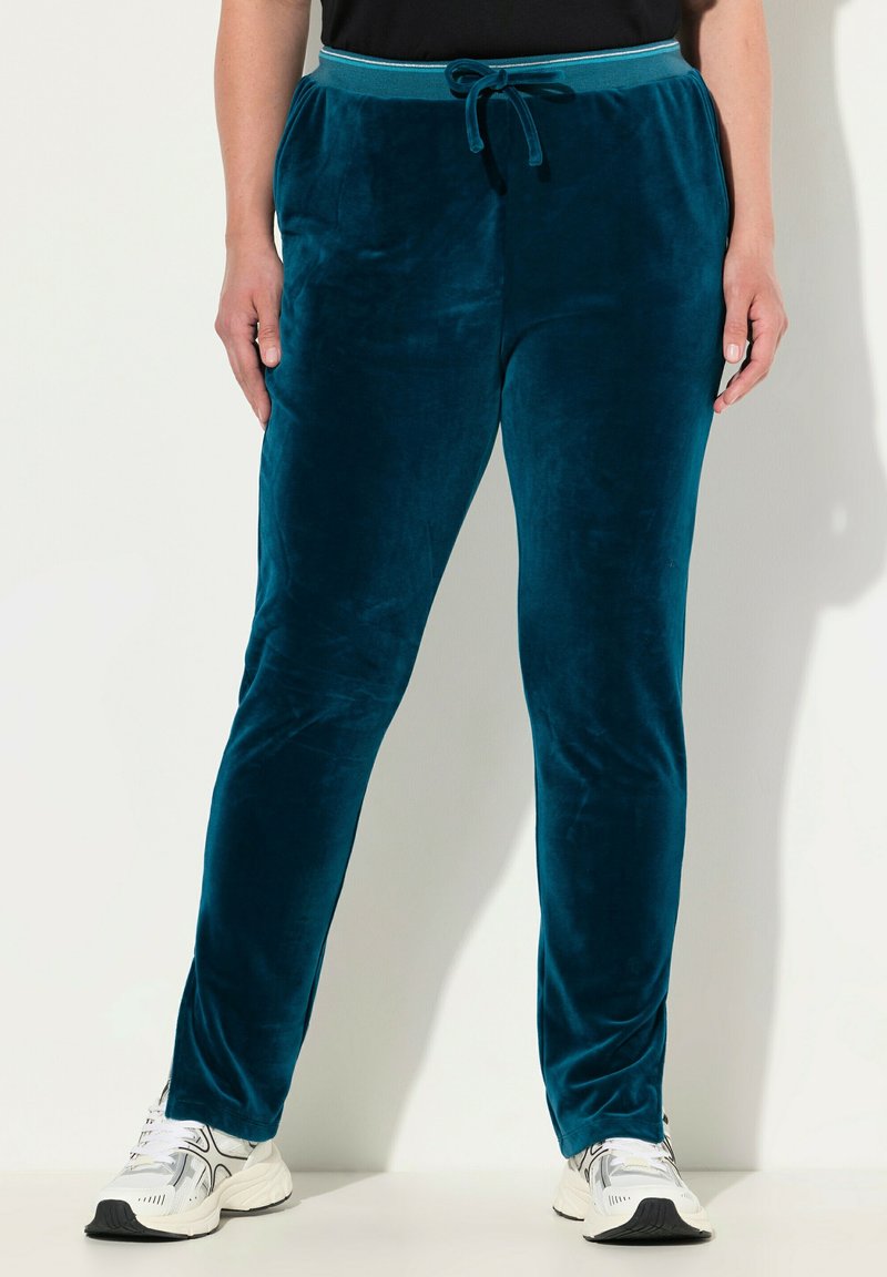 Teal fluwelen broek met een elastische tailleband en contrasterende blauwe bies. De stof heeft een gladde, zachte textuur. Gepaard met witte sportschoenen.