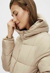 Beige dunjakke med quiltet tekstur, glidelås lukking og hette. Har ribbede mansjetter og en strømlinjeformet fasong, egnet for lagdeling.
