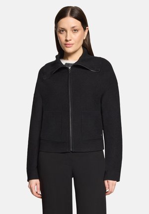 Frau mit glattem braunem Haar trägt eine schwarze Reißverschlussjacke mit zwei Vordertaschen und schwarze Hose vor einem schlichten hellen Hintergrund.