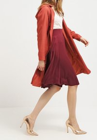 Rood geknoopte jas over een wit kanten top en een geplooide bordeauxrode rok. Beige hoge hakken maken de outfit af en benadrukken elegantie en beweging.