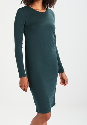 Femme portant une robe ajustée verte foncée à manches longues, longueur genou, debout devant un fond blanc uni.